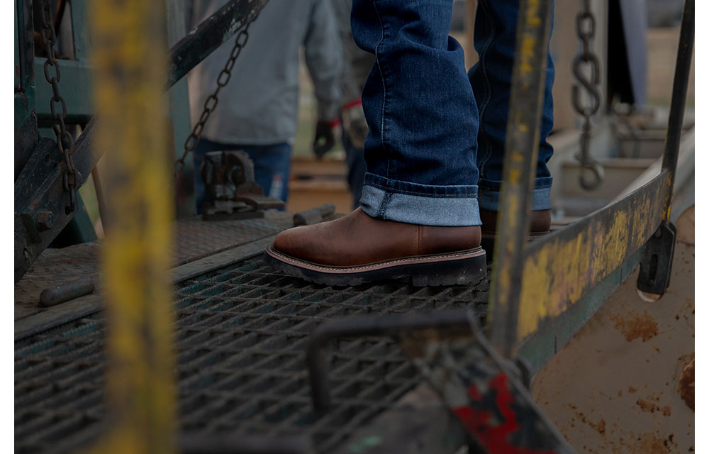 Un hombre que viste pantalones vaqueros con dobladillo y botas Homeland de color marrón está parado sobre una plataforma de rejilla de metal, con cadenas industriales y equipos visibles en el fondo.